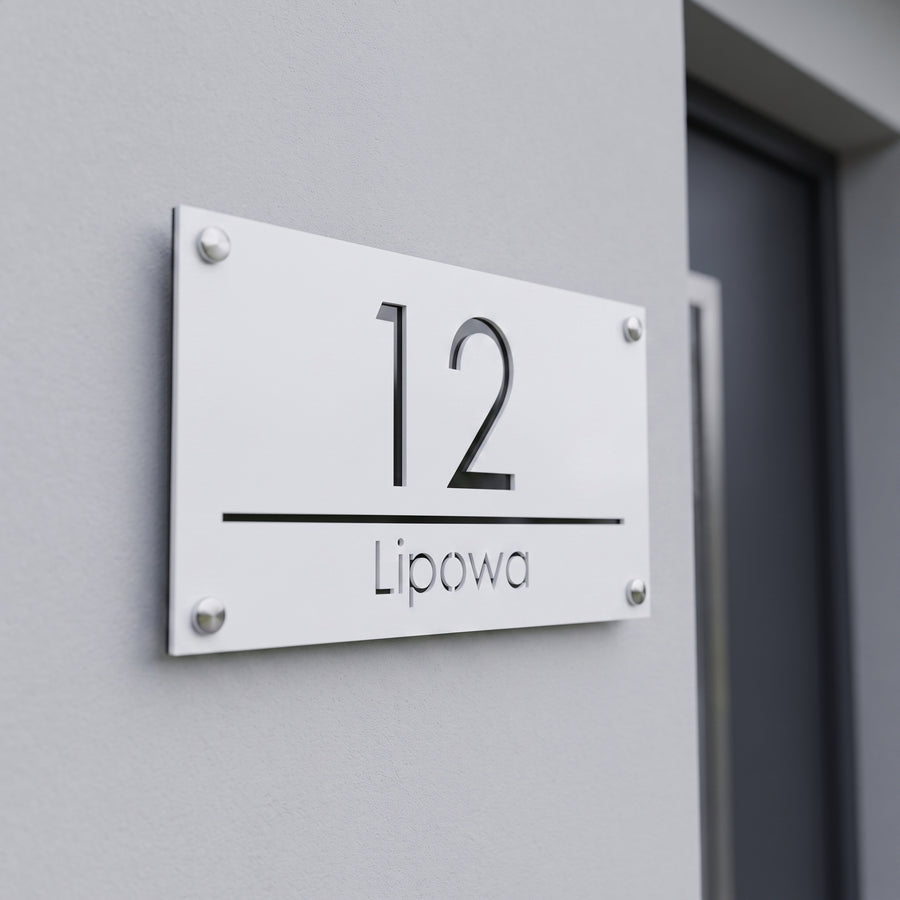Biała tabliczka adresowa 3D bez podświetlenia z numerem 12 i ulicą Lipowa na jasnoszarej elewacji w świetle dziennym